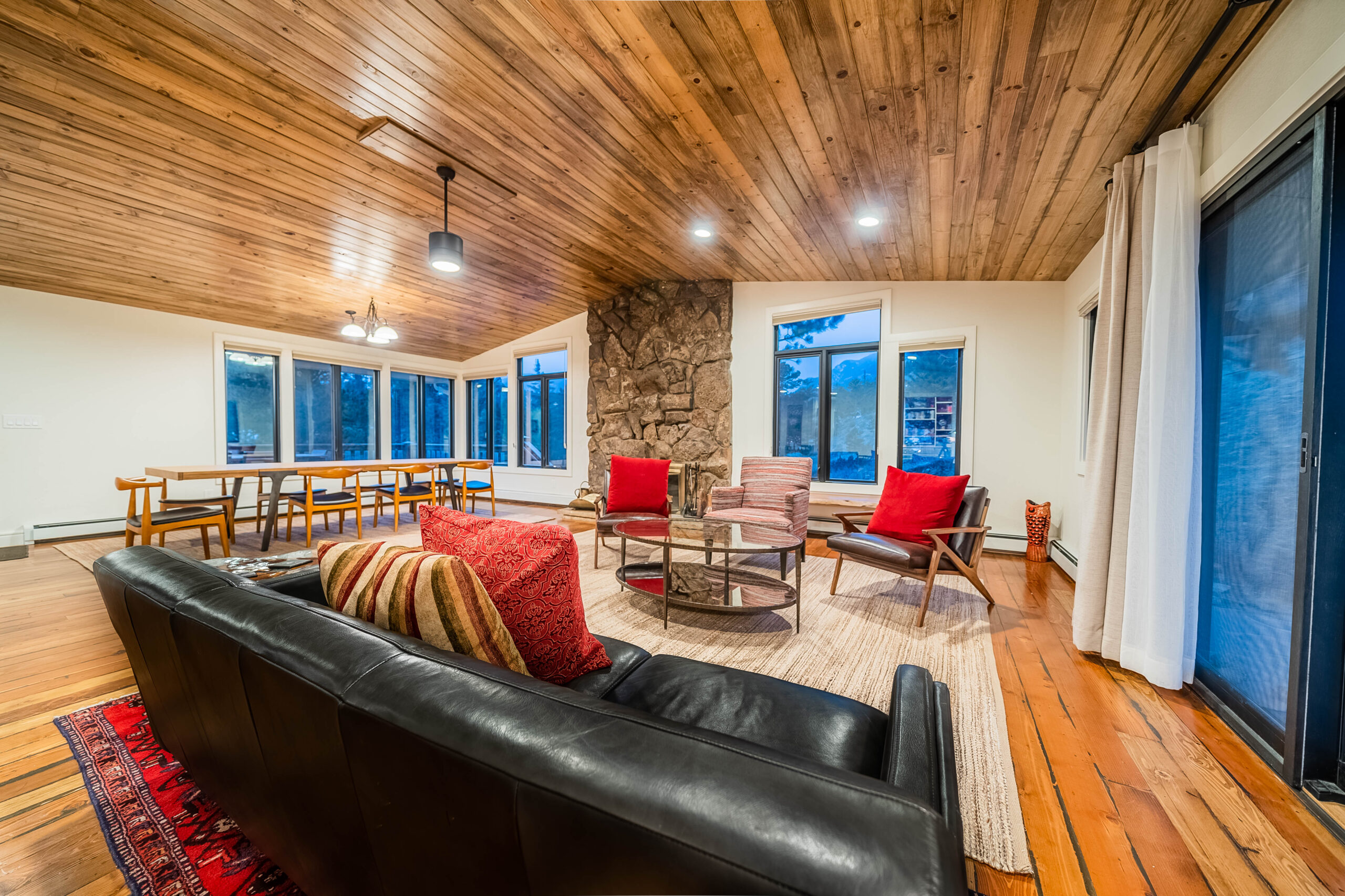A light-filled living room with a wood ceiling, dining table, and tall windows.