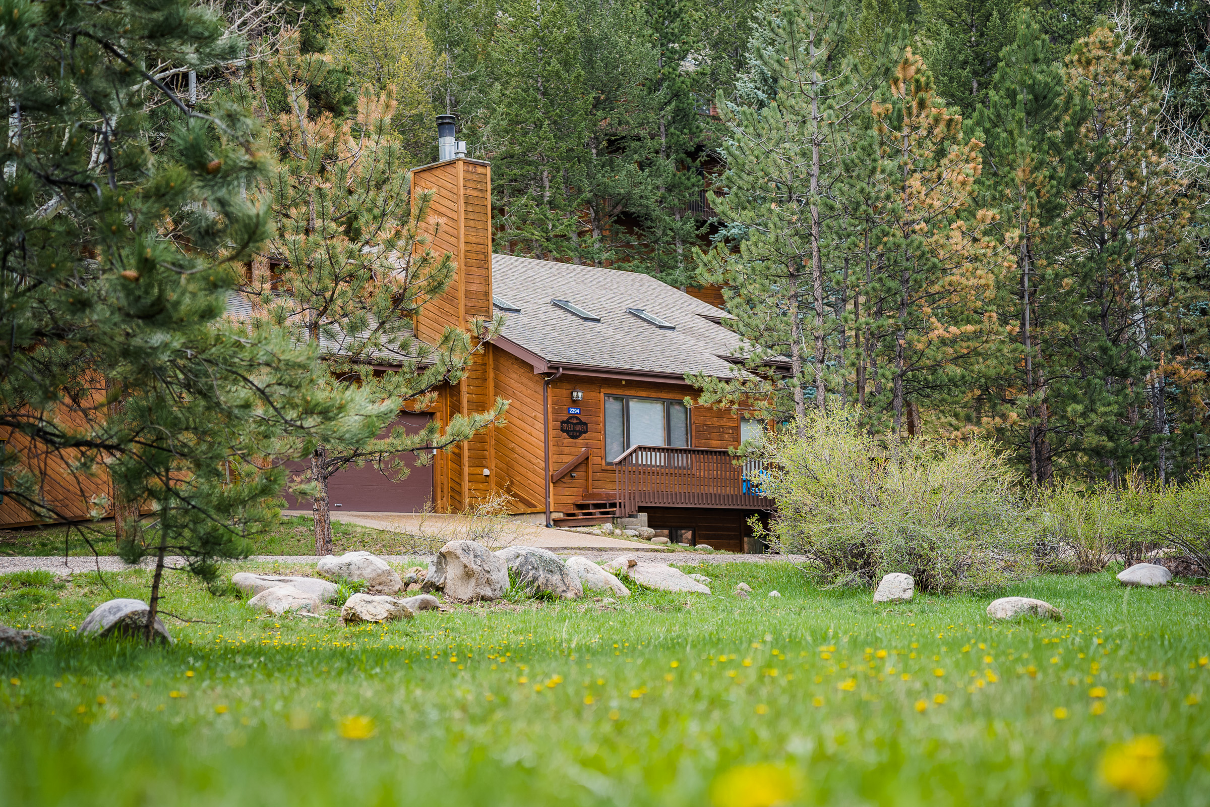 A cabin among green trees, with a lawn and boulders beside the porch.