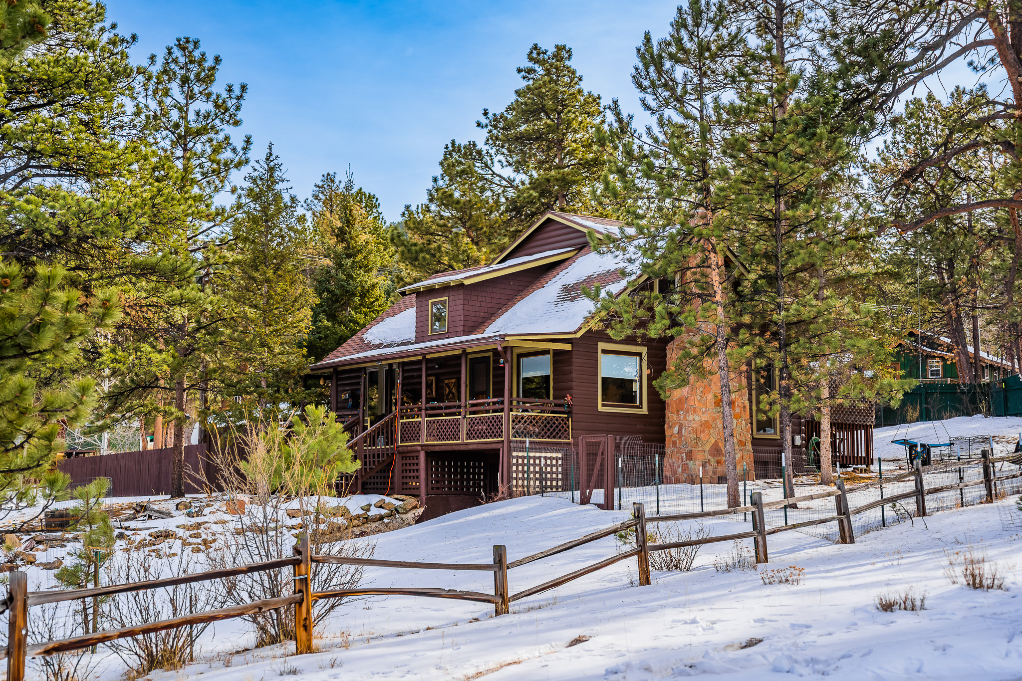 A wooden cabin with a stone chimney is surrounded by snow and pine trees. A picket fence lines the foreground. The sky is clear and blue.