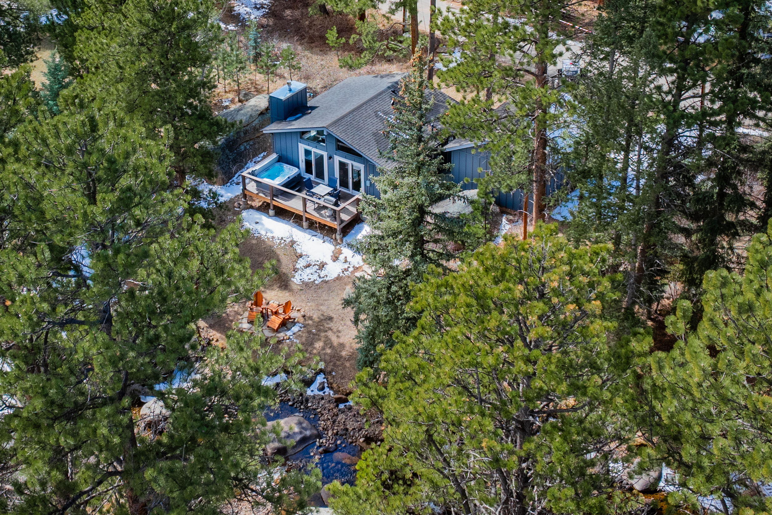 Aerial view of a cabin nestled among pine trees. The cabin has a grey roof and large windows. A wooden deck with a hot tub is visible. Snow patches are scattered on the ground and a small creek runs nearby.
