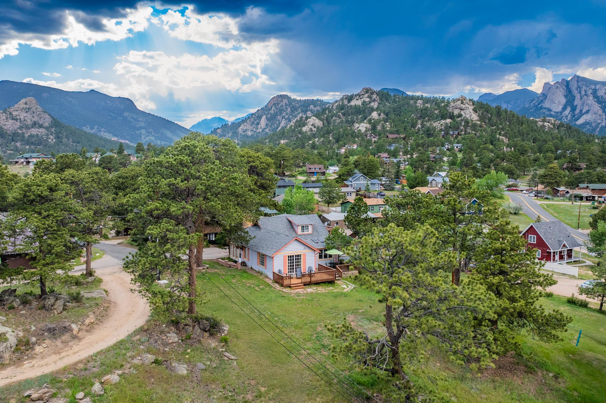 A small house with a porch is surrounded by trees and sits on a grassy lot with a dirt driveway. Other houses and buildings are spread throughout the valley, with forested mountains and a cloudy blue sky in the background.