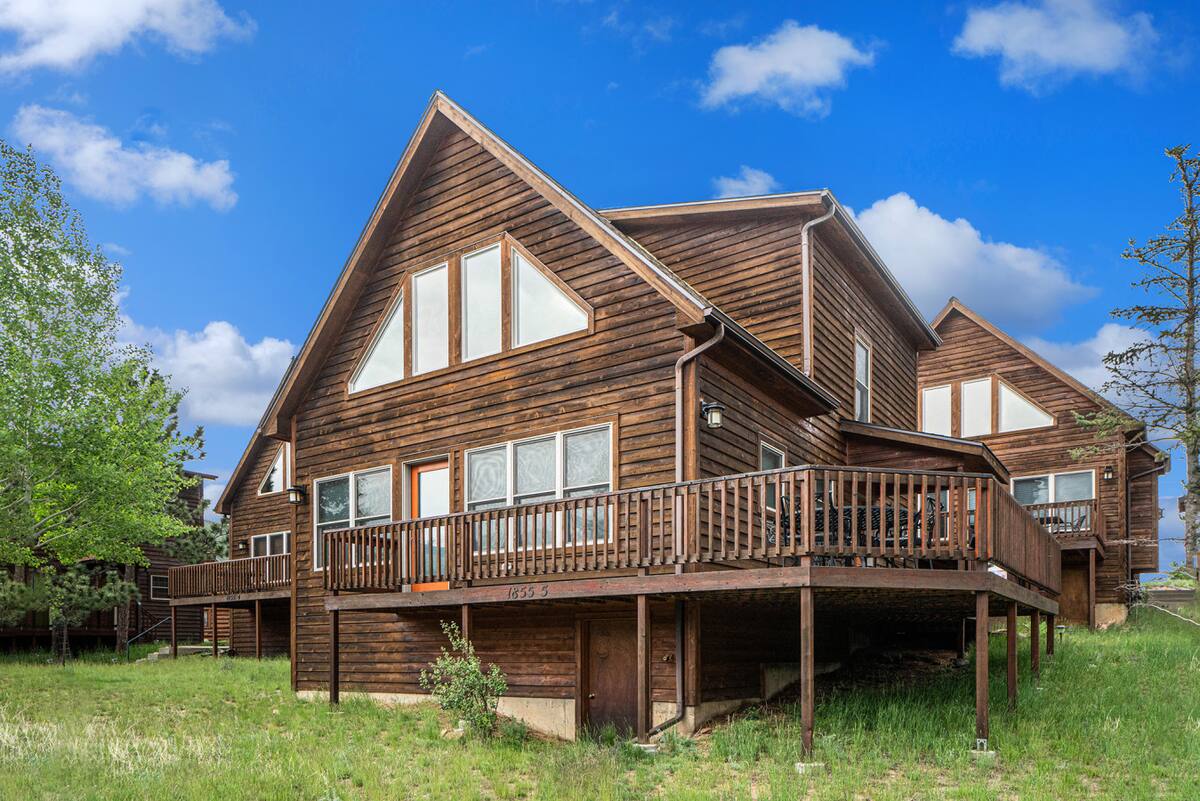 A large wooden house with an A-frame roof, multiple triangular and rectangular windows, a wide wraparound deck, and elevated foundation, set on a grassy lot under a blue sky with scattered clouds.