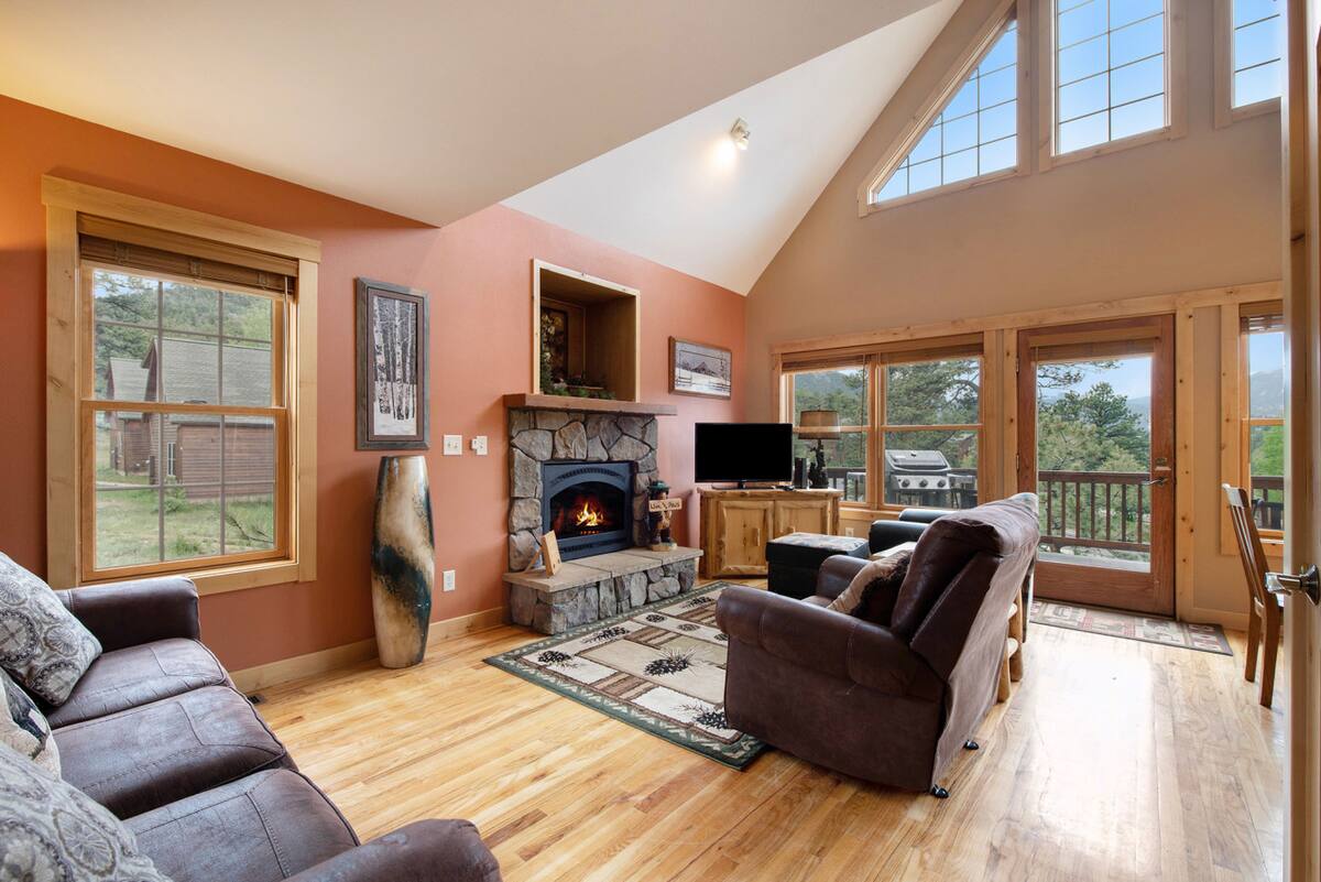 A living room with wooden floors, two brown sofas, a stone fireplace with a lit fire, a TV on a stand, large windows, and glass doors leading to a balcony with trees visible outside.
