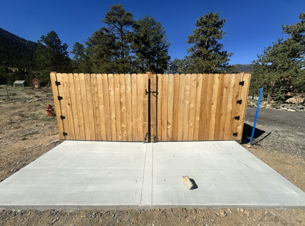 A wooden double gate stands closed on a new concrete pad, with a metal latch in the center. There are trees, a blue sky, rocky ground, and a fire hydrant in the background. A small rock lies on the concrete.