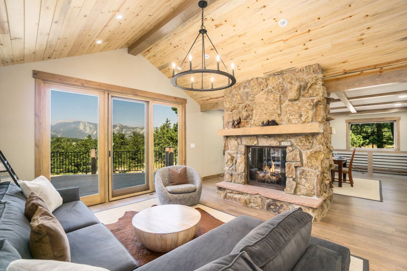 A cozy living room with a stone fireplace, gray sofa, round coffee table, and a single armchair. Large glass doors open to a deck with mountain views. The ceiling and floor are wood, and a chandelier hangs above.