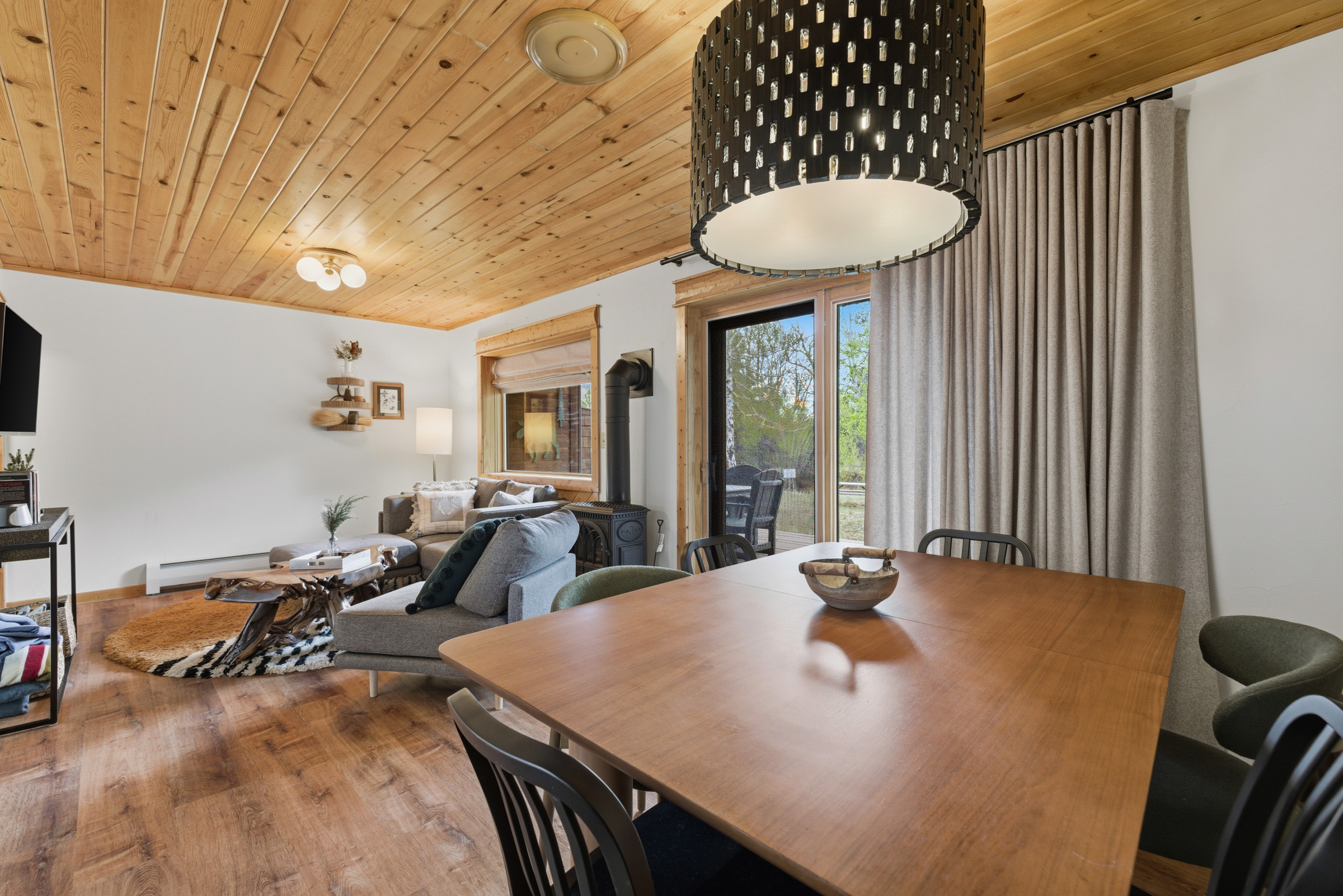 A modern living and dining area with wood floors and a wood-paneled ceiling. The space has a dining table, black chairs, a gray sectional sofa, a unique wood coffee table, and large windows with curtains looking outside.